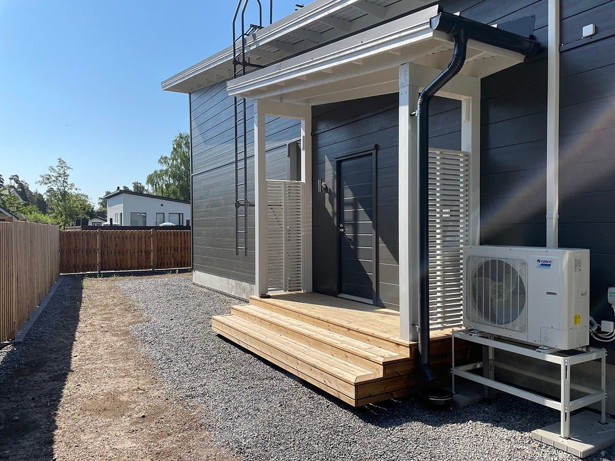 Yksikerroksinen harmaa talo valkoisin reunuksin, pieni kuisti ja harjakatto, sorapolun ja harvojen puiden ympäröimänä sinisen taivaan alla. Vasemmalle puolelle on asennettu lämpöpumppu.