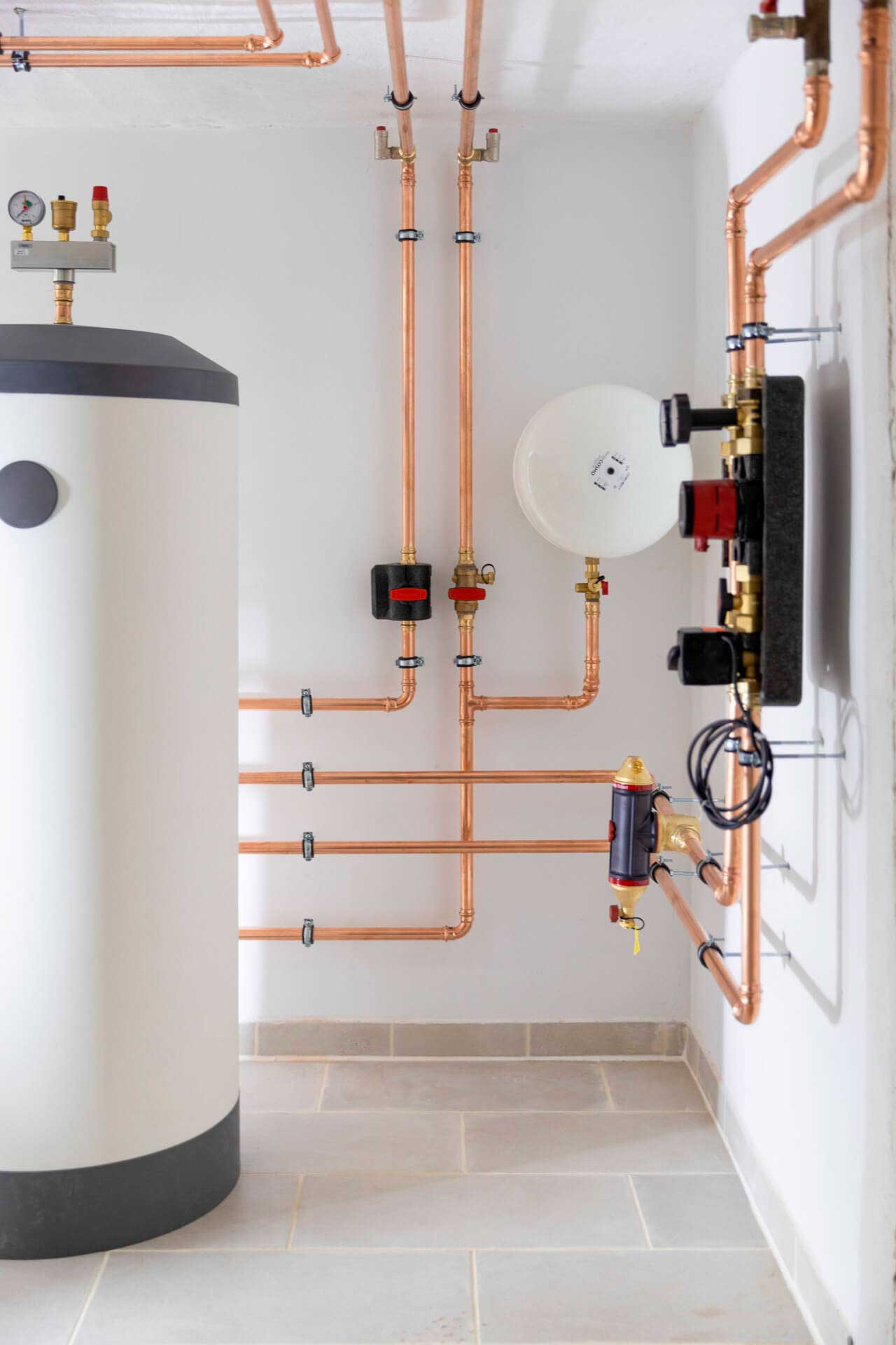 Moderni kattilahuone, jossa on kupariputkia, suuri sylinterimäinen lämminvesivaraaja, mittareita ja ohjauspaneeli vaaleasävyisellä laattalattialla.