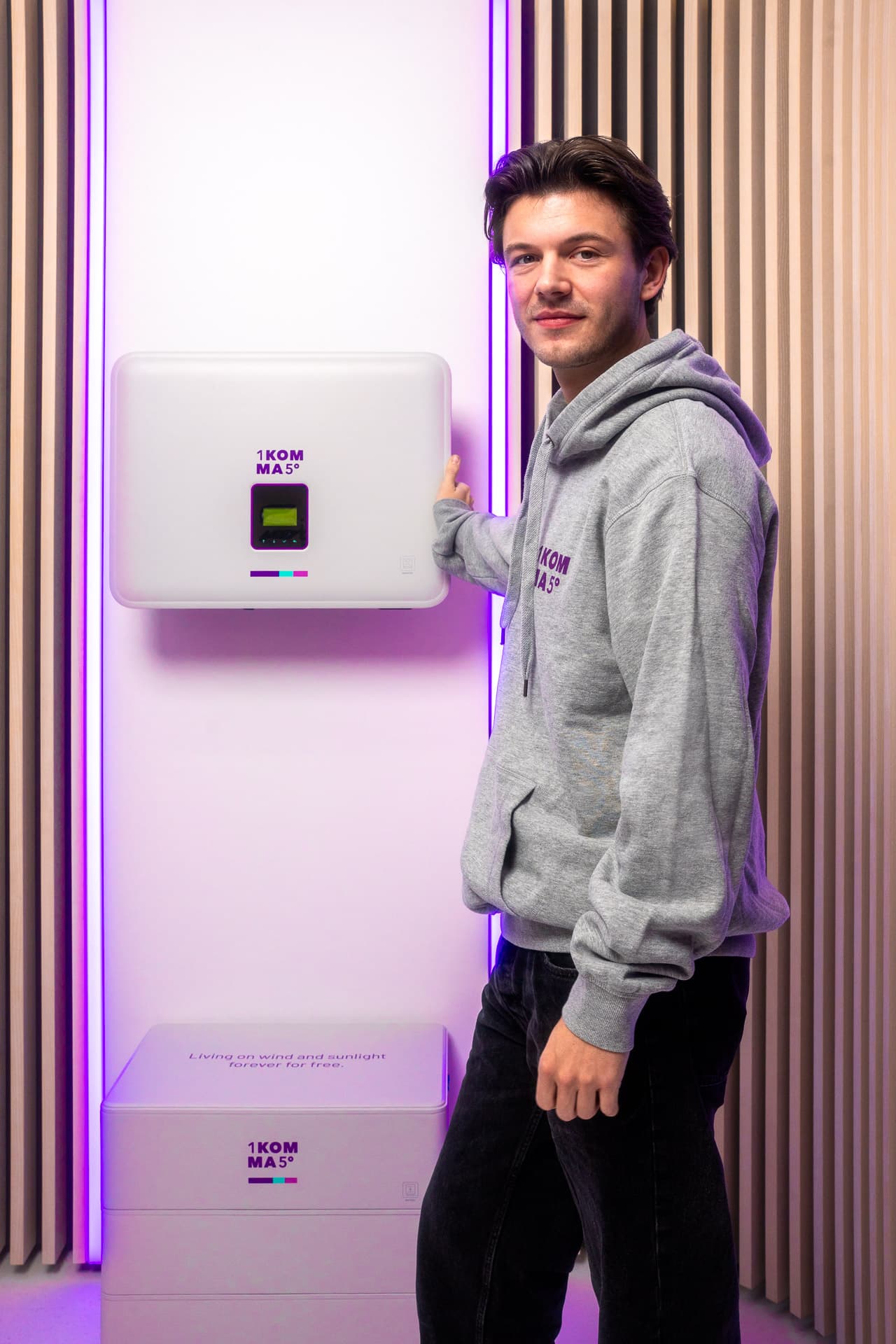Henkilö harmaassa hupparissa seisoo seinälle asennetun invertterilaitteen vieressä, jossa on violetti taustavalaistus, ja koskettaa sen sivua.