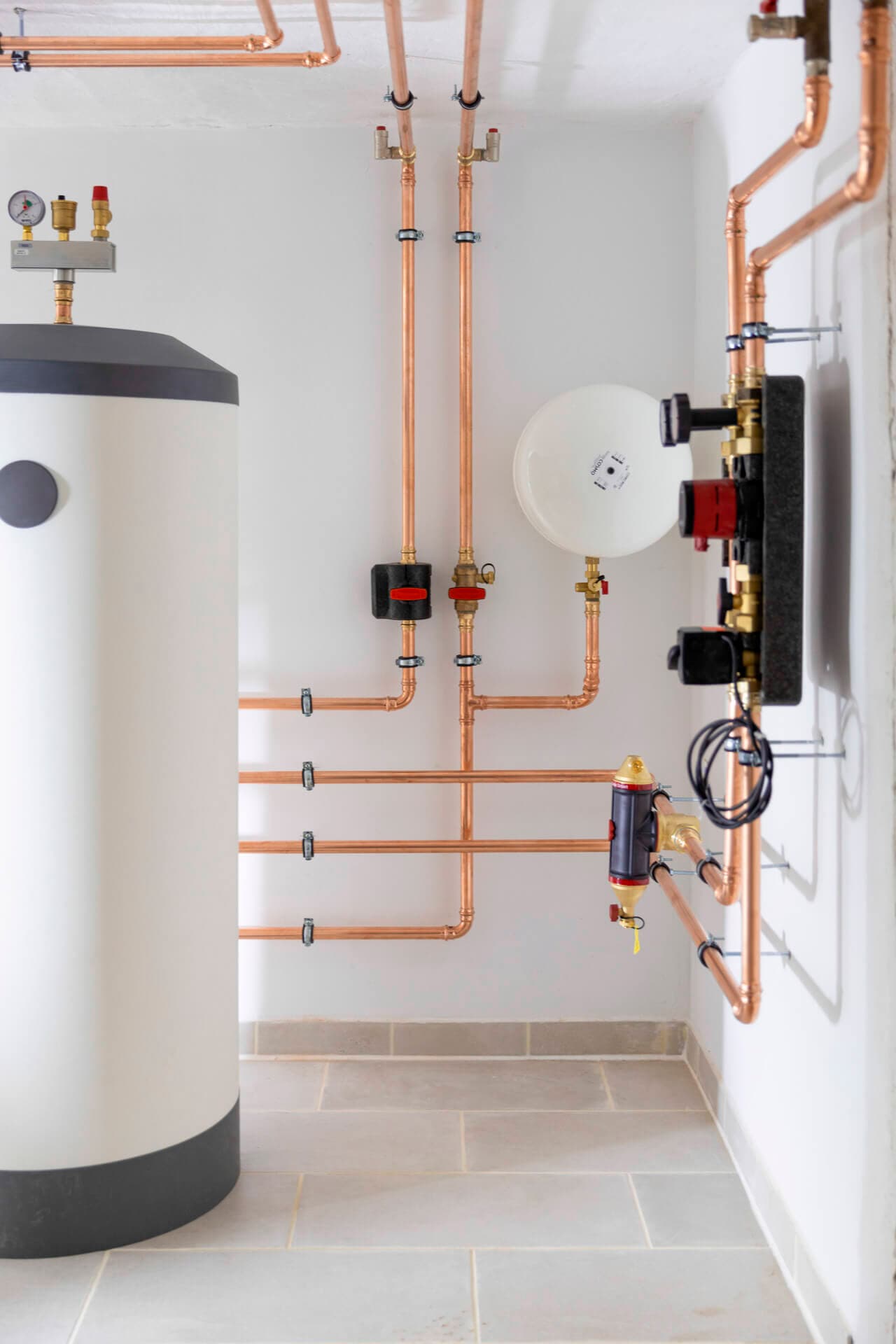 Moderni kattilahuone, jossa on sylinterimäinen lämminvesivaraaja ja kupariputkia valkoiselle seinälle asennettuna.