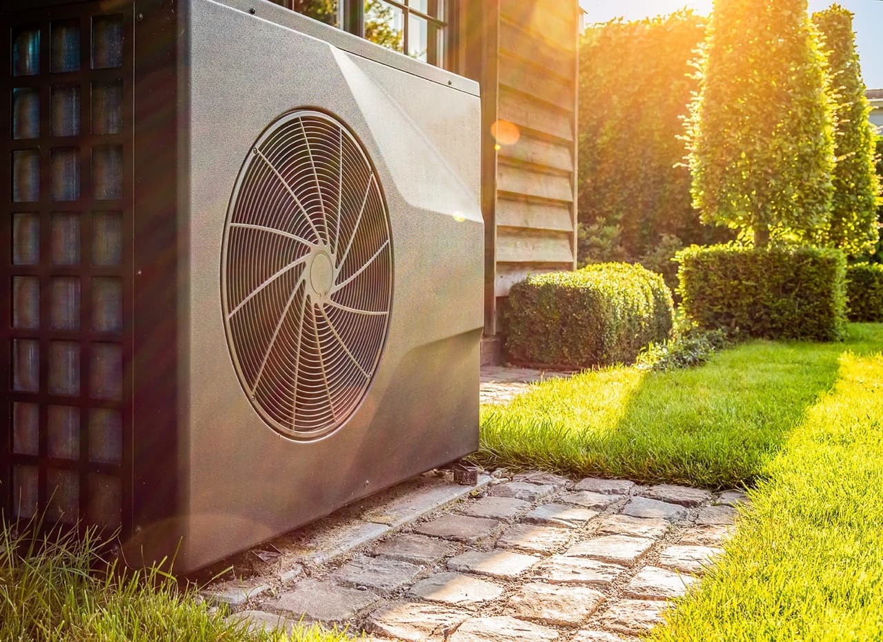 Ulkolämpöpumppuyksikkö asennettuna talon viereen, vihreän nurmikon ja siististi leikattujen pensasaitojen ympäröimänä kirkkaassa auringonvalossa.