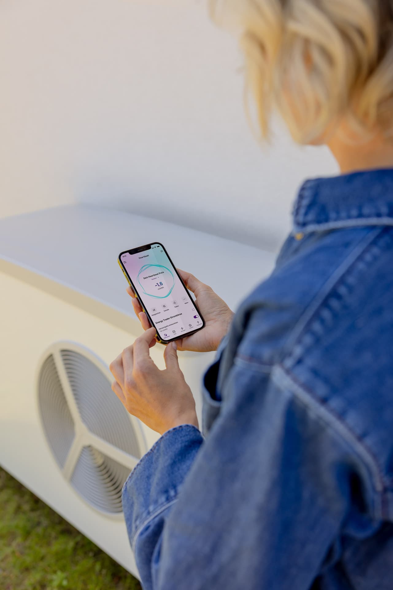Henkilö farkkutakissa käyttää älypuhelinta ohjatakseen valkoista ulkona olevaa ilmastointilaitetta.
