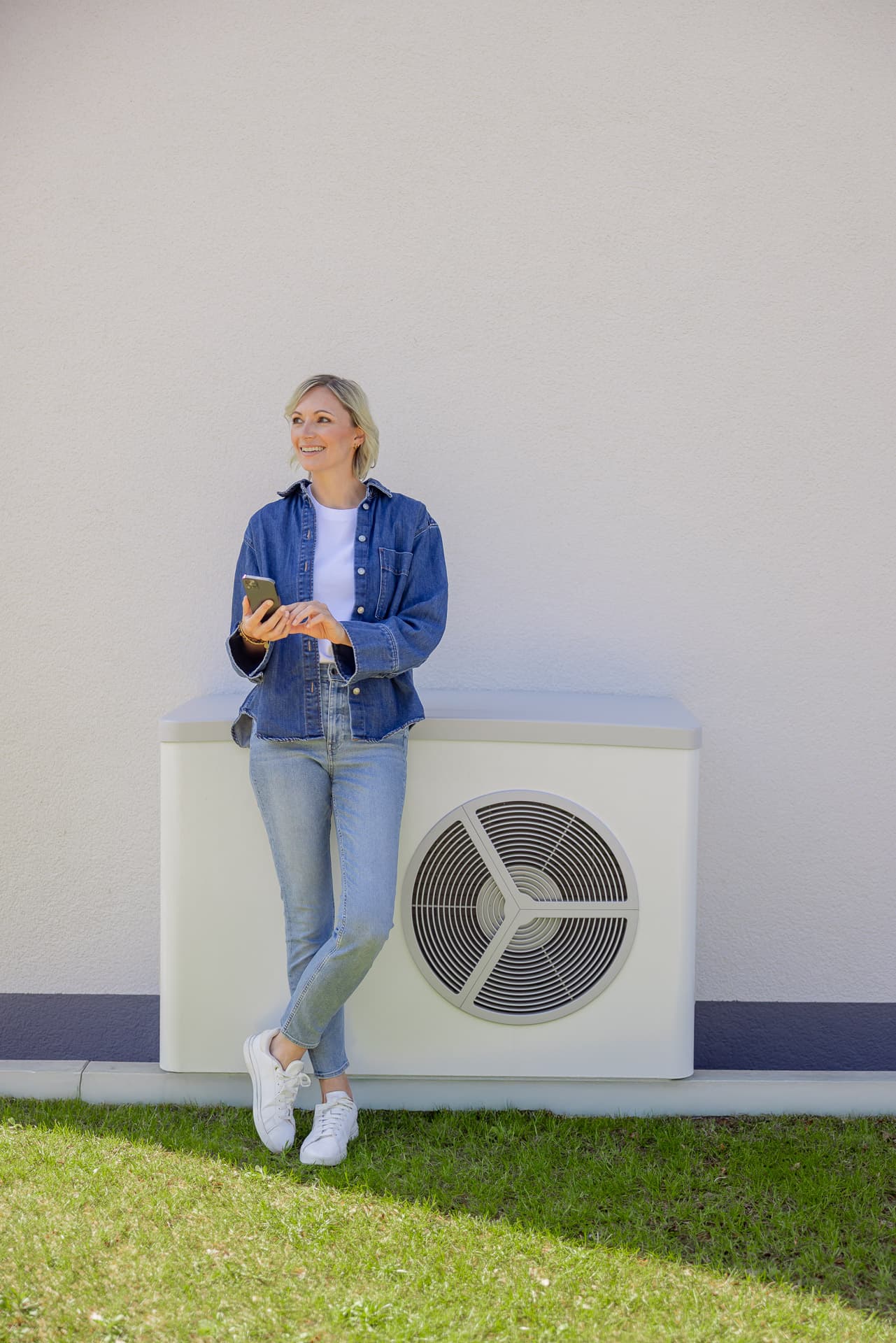 Nainen farkkuasussa nojaa valkoiseen ulkolämpöpumppuyksikköön, pitelee puhelinta ja seisoo nurmikolla vaalean seinän edessä.
