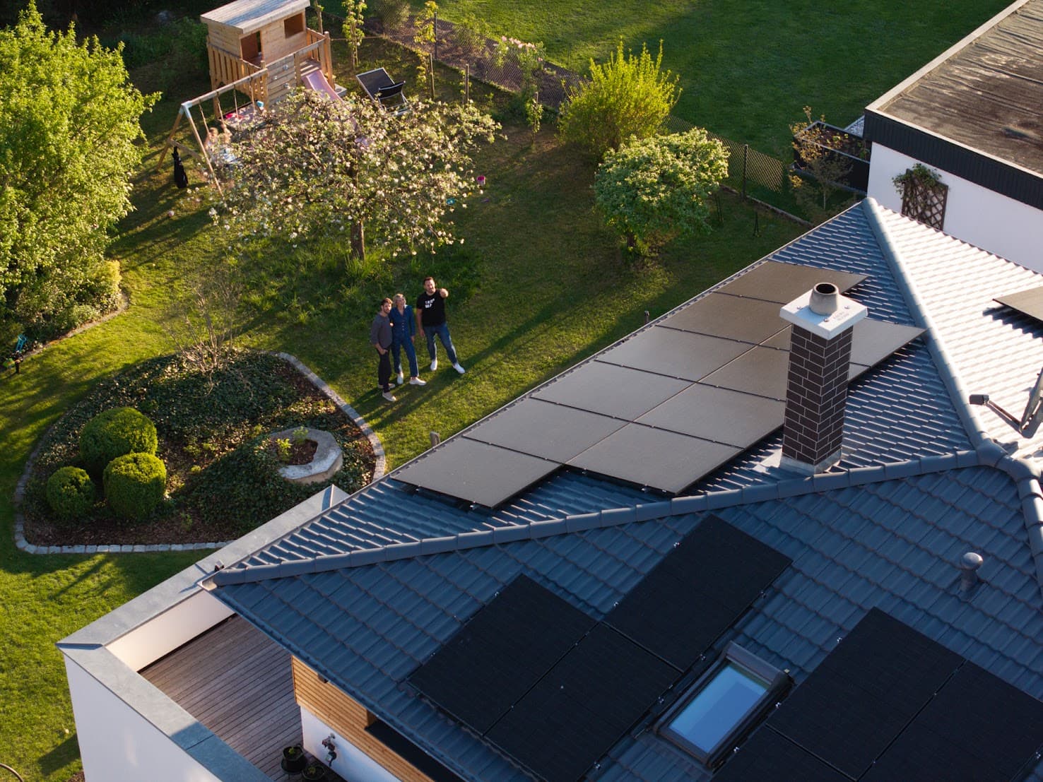 Ilmakuva talosta, jonka katolla on aurinkopaneelit, puutarhan ympäröimänä, jossa on puita ja pieni leikkialue. Kolme ihmistä seisoo puutarhassa.