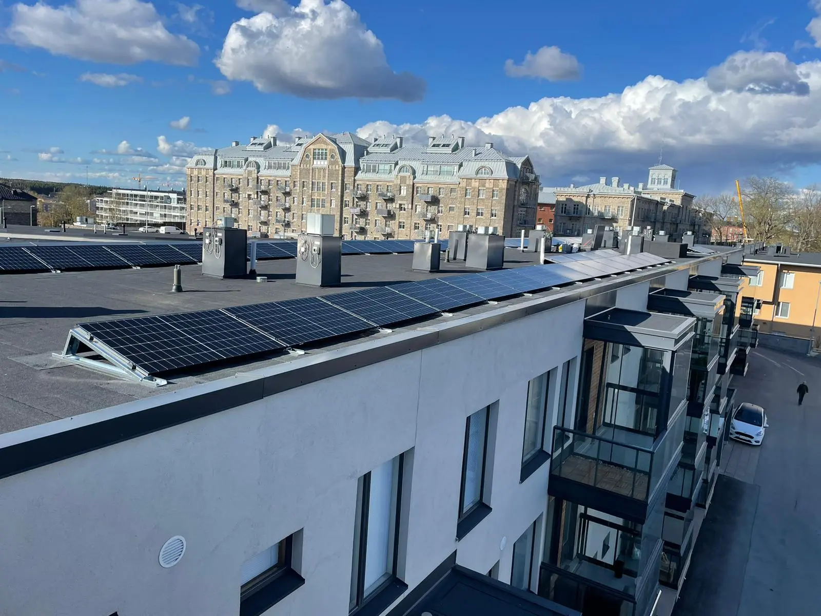 Aurinkopaneelit nykyaikaisten rakennusten katoilla sinisen, pilvisen taivaan alla, taustalla historiallista arkkitehtuuria.