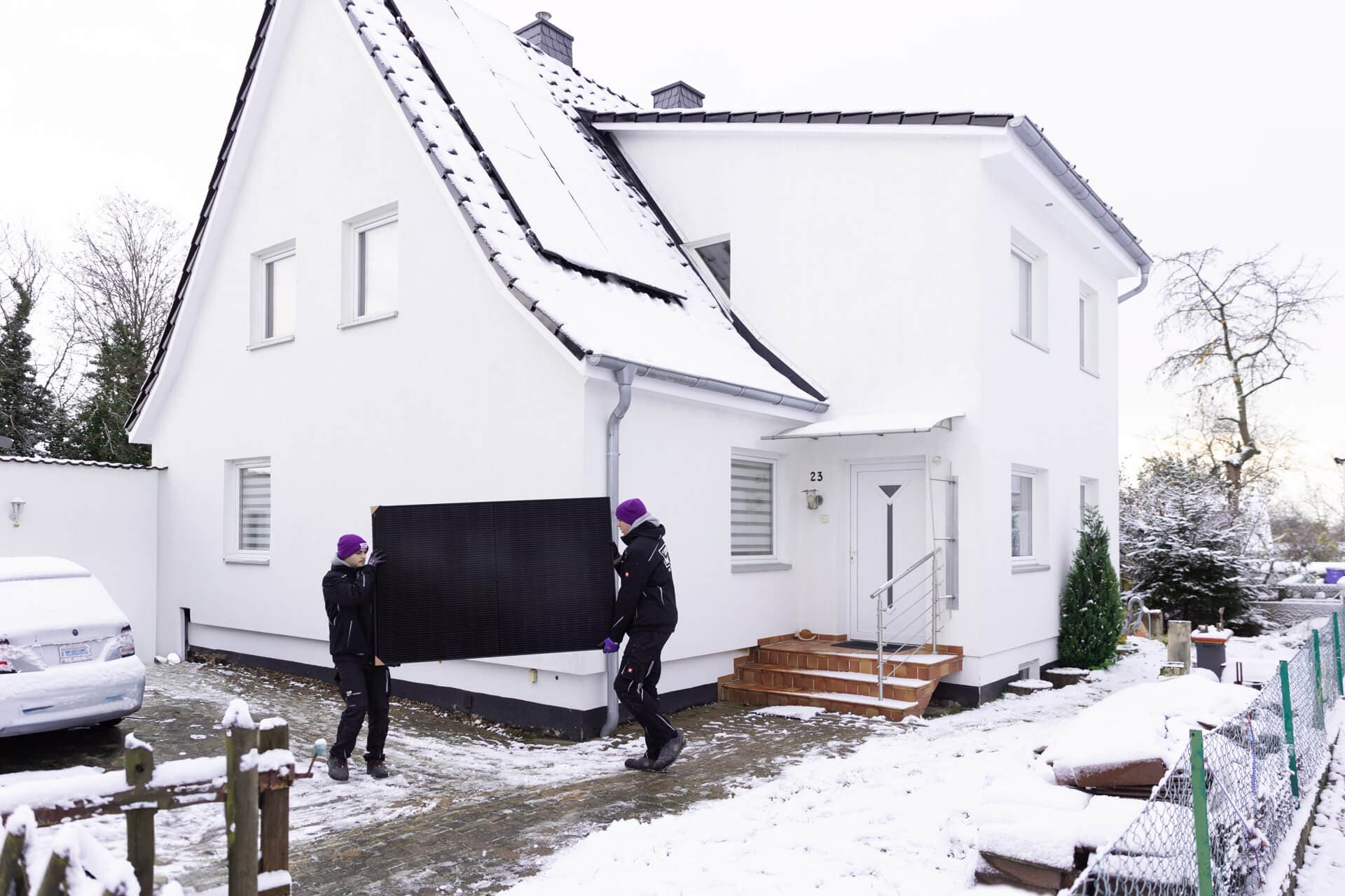 Kaksi ihmistä kantaa aurinkopaneelia lumen peittämän valkoisen jyrkkäkattoisen talon ulkopuolella.