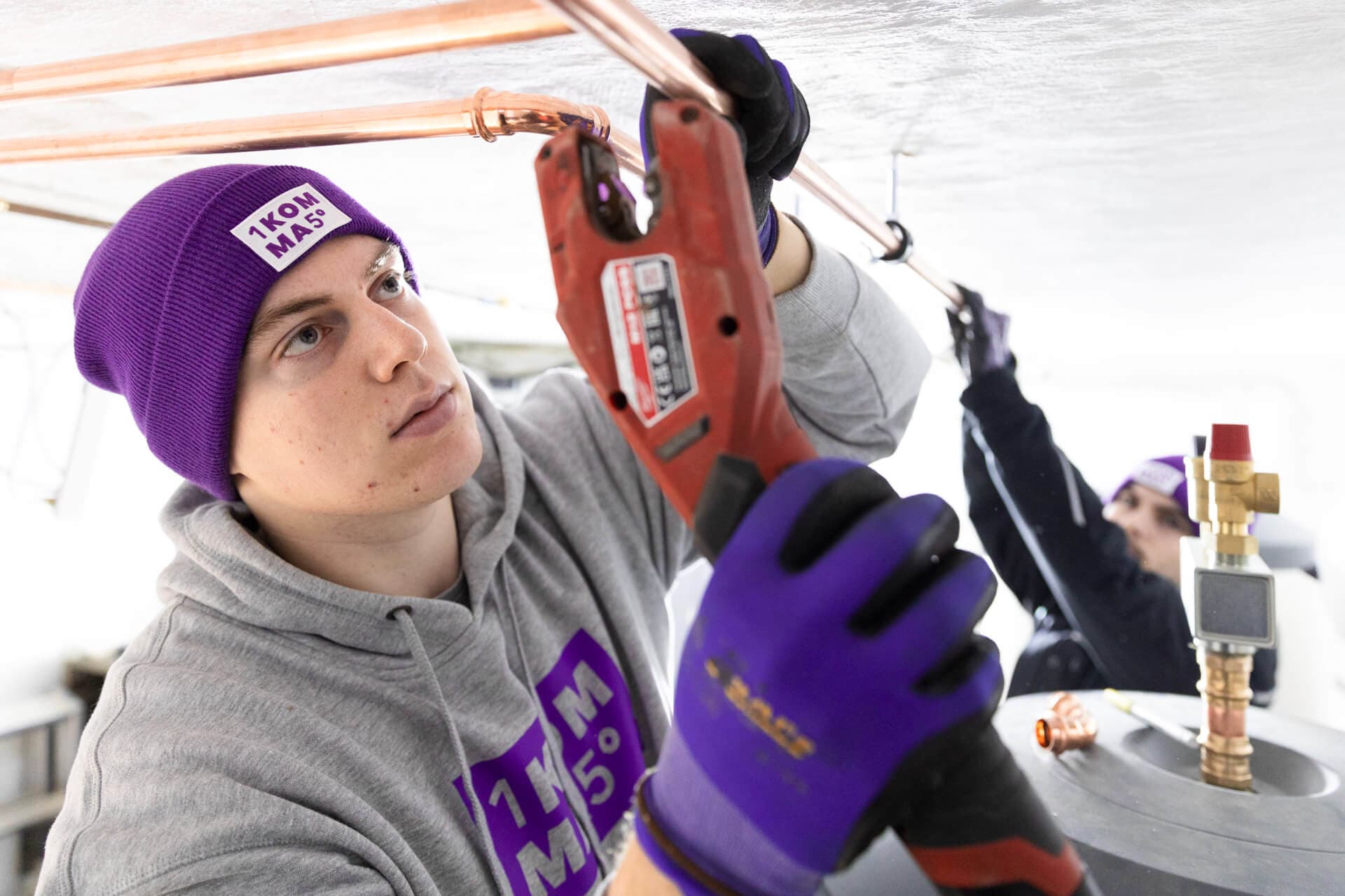 Henkilö violetissa pipossa ja käsineissä työskentelee kupariputkien parissa työkalua käyttäen. Toinen henkilö näkyy taustalla.