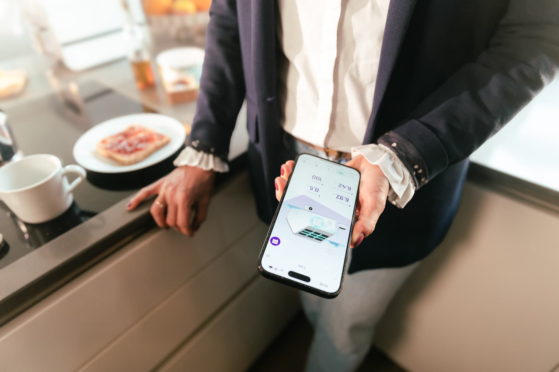 Henkilö pitelee älypuhelinta, jonka näytöllä on sovellus, seisten keittiötason ääressä, jossa on lautasellinen vohveleita ja kuppi.