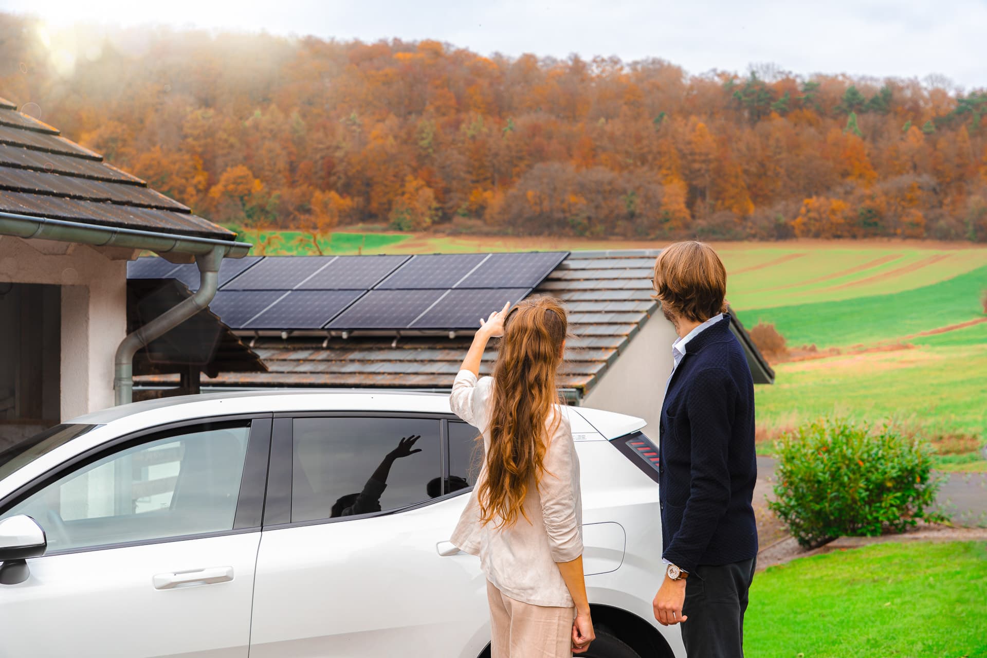 Pariskunta seisoo sähköauton vieressä ja osoittaa talon katolla olevia aurinkopaneeleja syksyisten puiden ja peltojen taustalla.