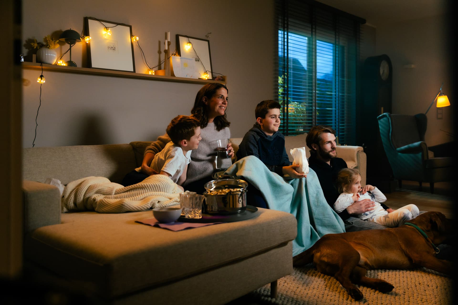 Perhe istuu sohvalla vilttien alla katsoen televisiota. Koira makaa lattialla. Huone on hämärästi valaistu koristevaloin.