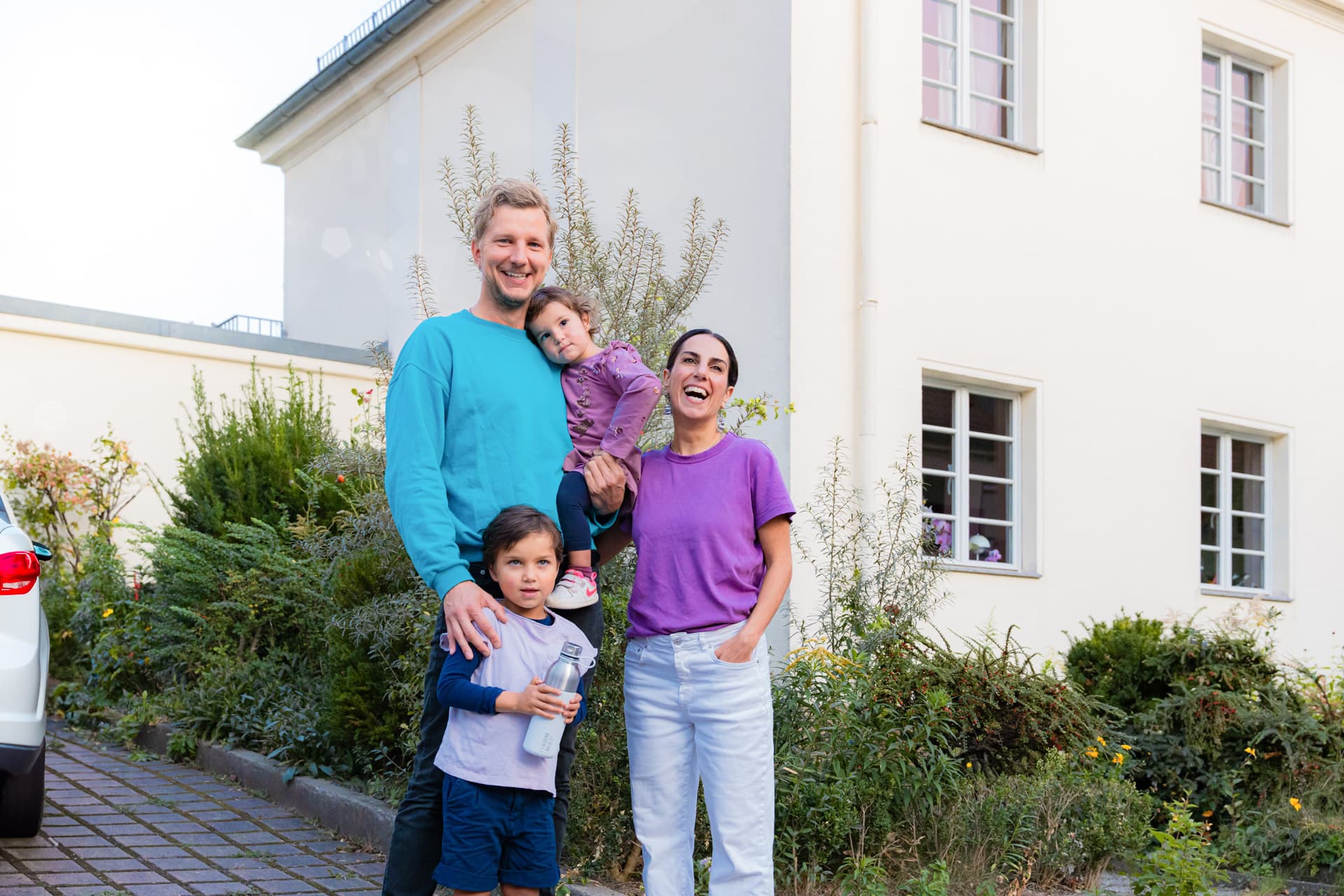 Hymyilevä nelihenkinen perhe seisoo talon edessä. Mies pitelee lasta sylissään, ja nainen seisoo heidän vieressään toinen lapsi edessään.