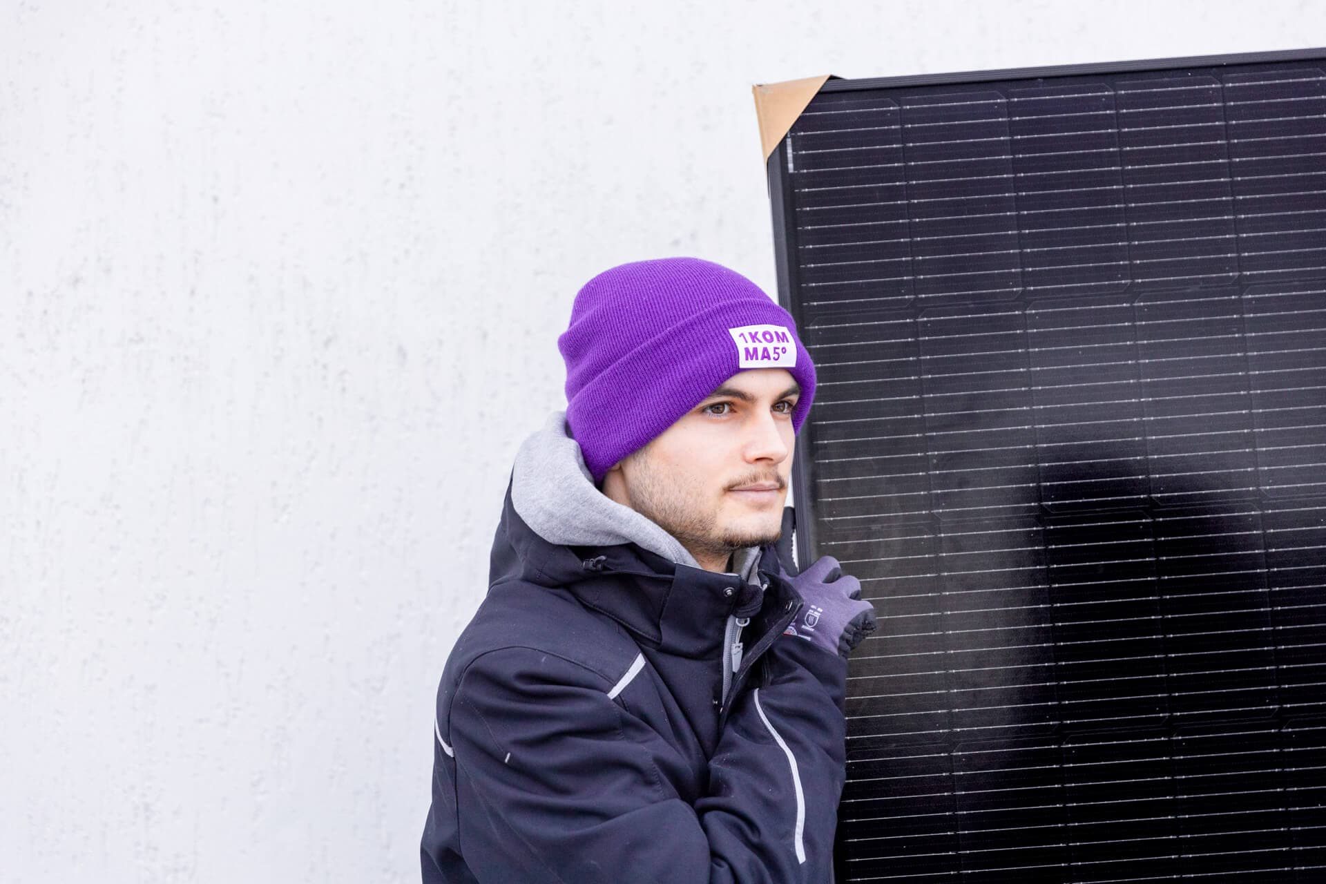 Henkilö violetissa pipossa ja tummassa takissa pitelee suurta aurinkopaneelia vaaleaa seinää vasten.