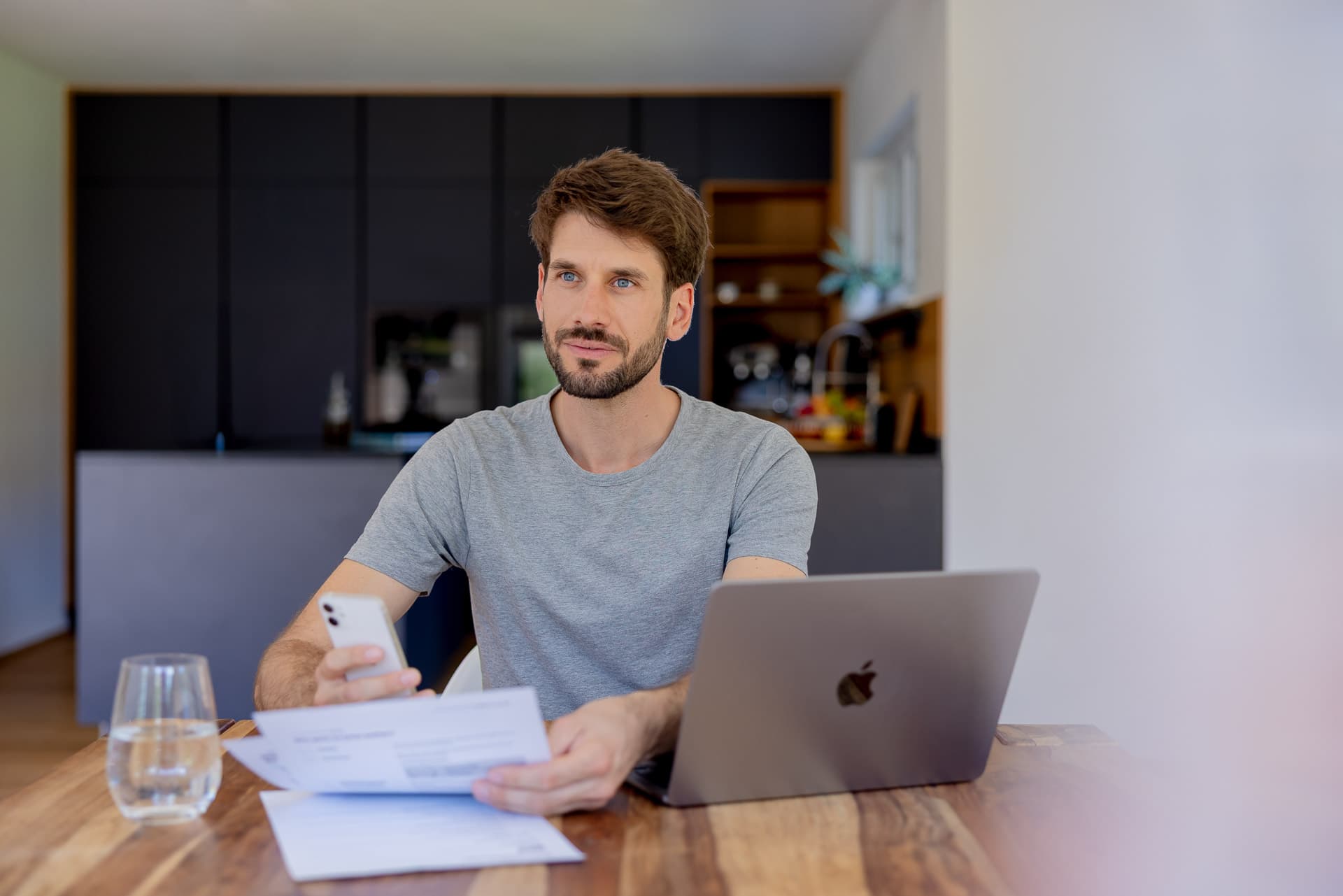 Mies istuu pöydän ääressä kannettavan tietokoneen kanssa, pitäen kädessään älypuhelinta ja asiakirjoja, modernissa keittiöympäristössä.