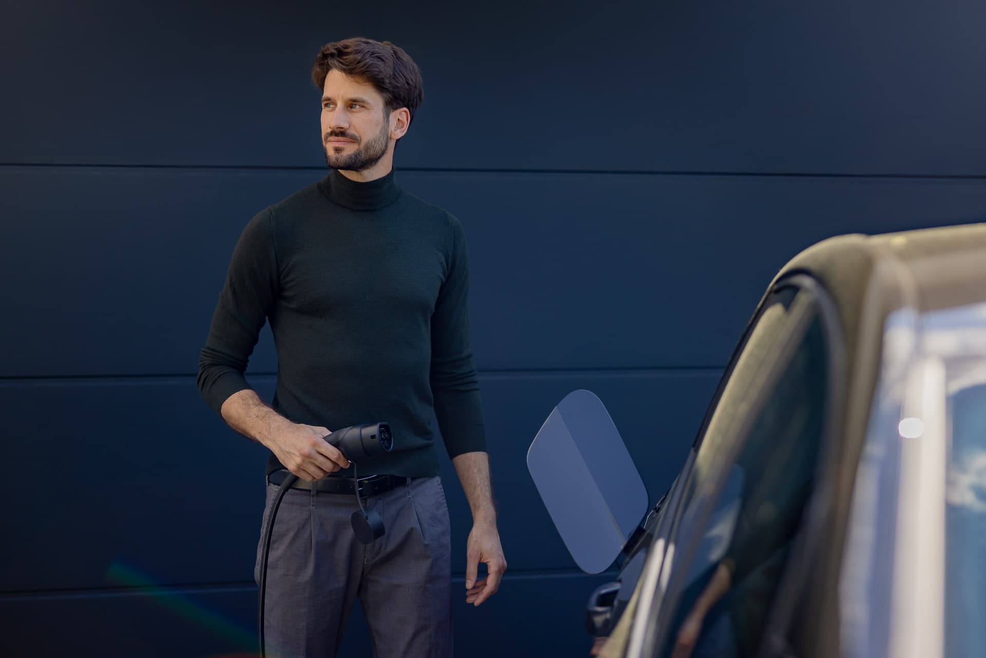 Mies mustassa poolopaidassa pitelee sähköauton laturia seisten auton vieressä tumman seinän edessä.