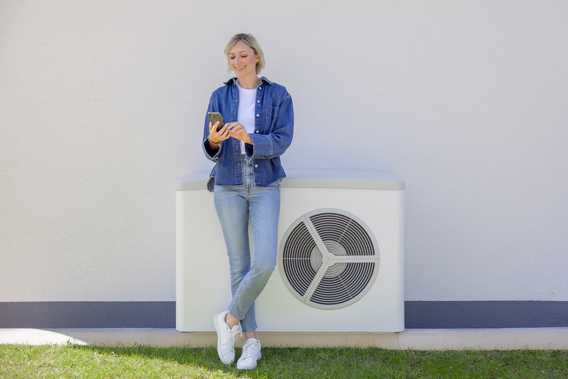 Nainen farkkuasussa nojaa ulkona olevaan ilmastointilaitteeseen ja katsoo puhelintaan, taustalla valkoinen seinä ja nurmikko.