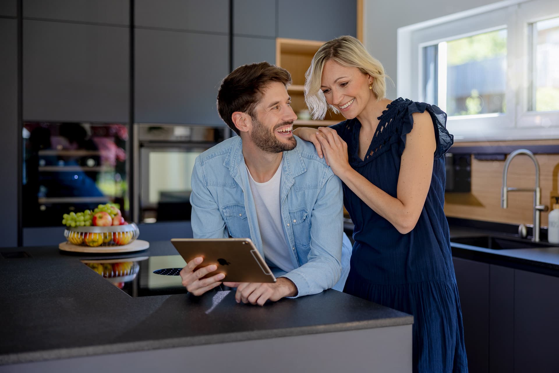 Hymyilevä pariskunta modernissa keittiössä, mies pitelee tablettia ja nainen nojaa hänen olkaansa vasten. Tiskillä on hedelmätarjotin.