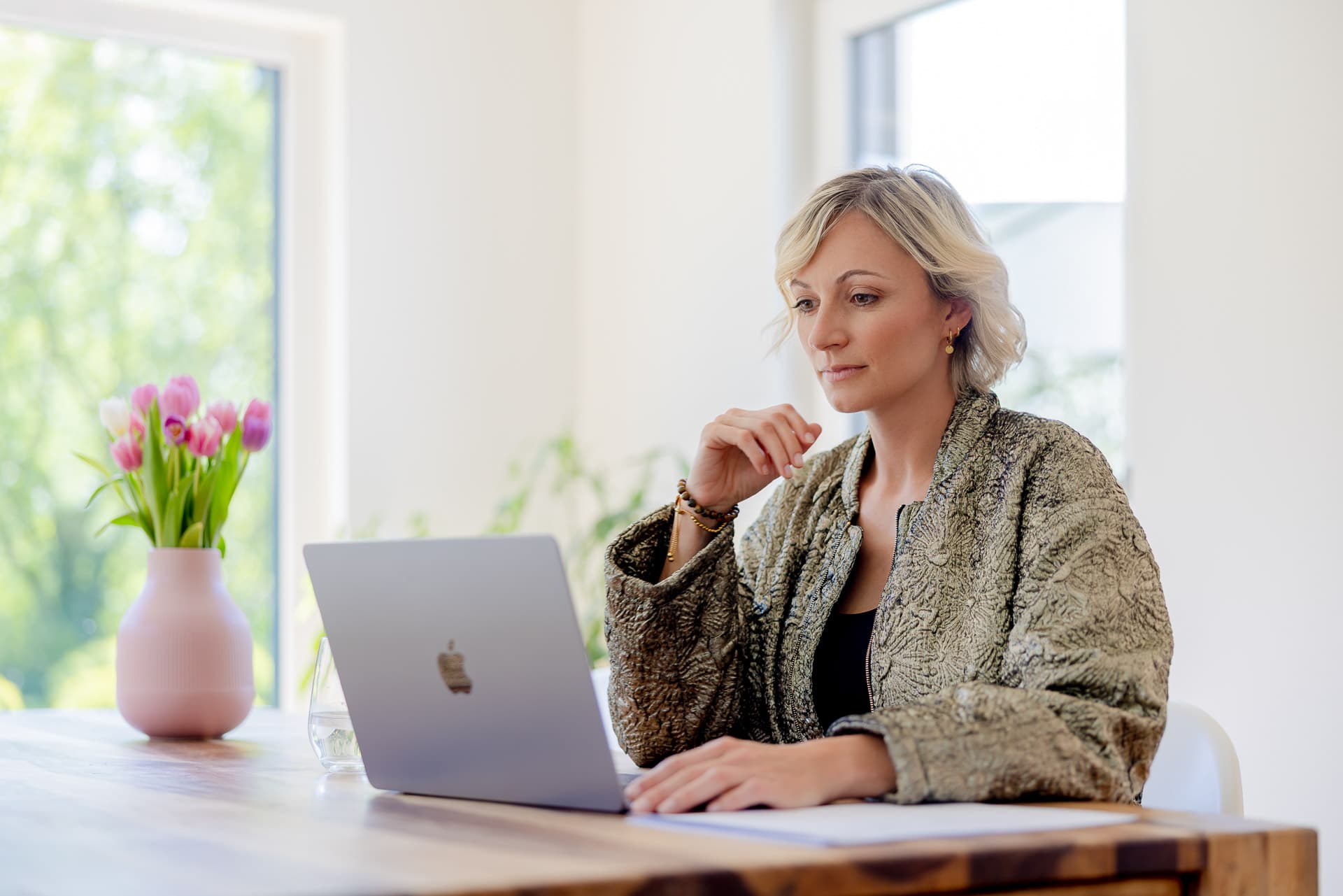 Nainen, jolla on lyhyet vaaleat hiukset, työskentelee kannettavalla tietokoneella puupöydän ääressä, lähellä maljakossa olevia vaaleanpunaisia tulppaaneja, valoisassa huoneessa.