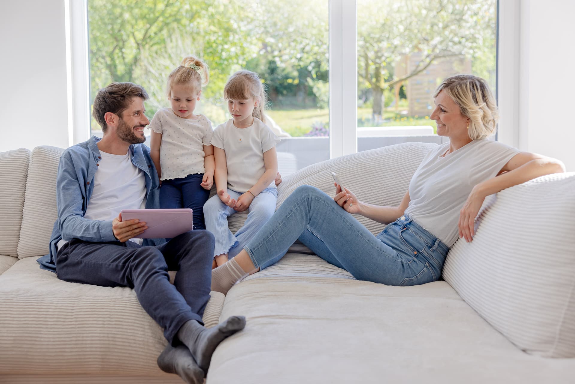 Neljän hengen perhe istuu sohvalla valoisassa huoneessa. Isä pitelee tablettia, äidillä on puhelin ja kaksi lasta istuu heidän välissään.