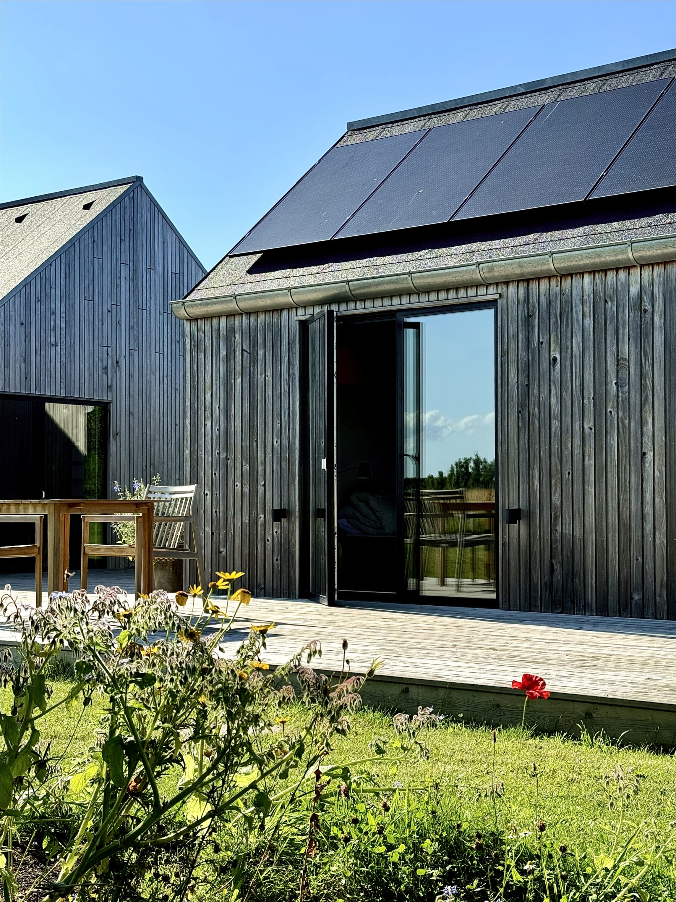 Moderni puutalo, jossa on aurinkopaneelit, lasiovi ja terassi pöytineen ja tuoleineen, nurmikon ja luonnonkukkien ympäröimänä kirkkaan taivaan alla.