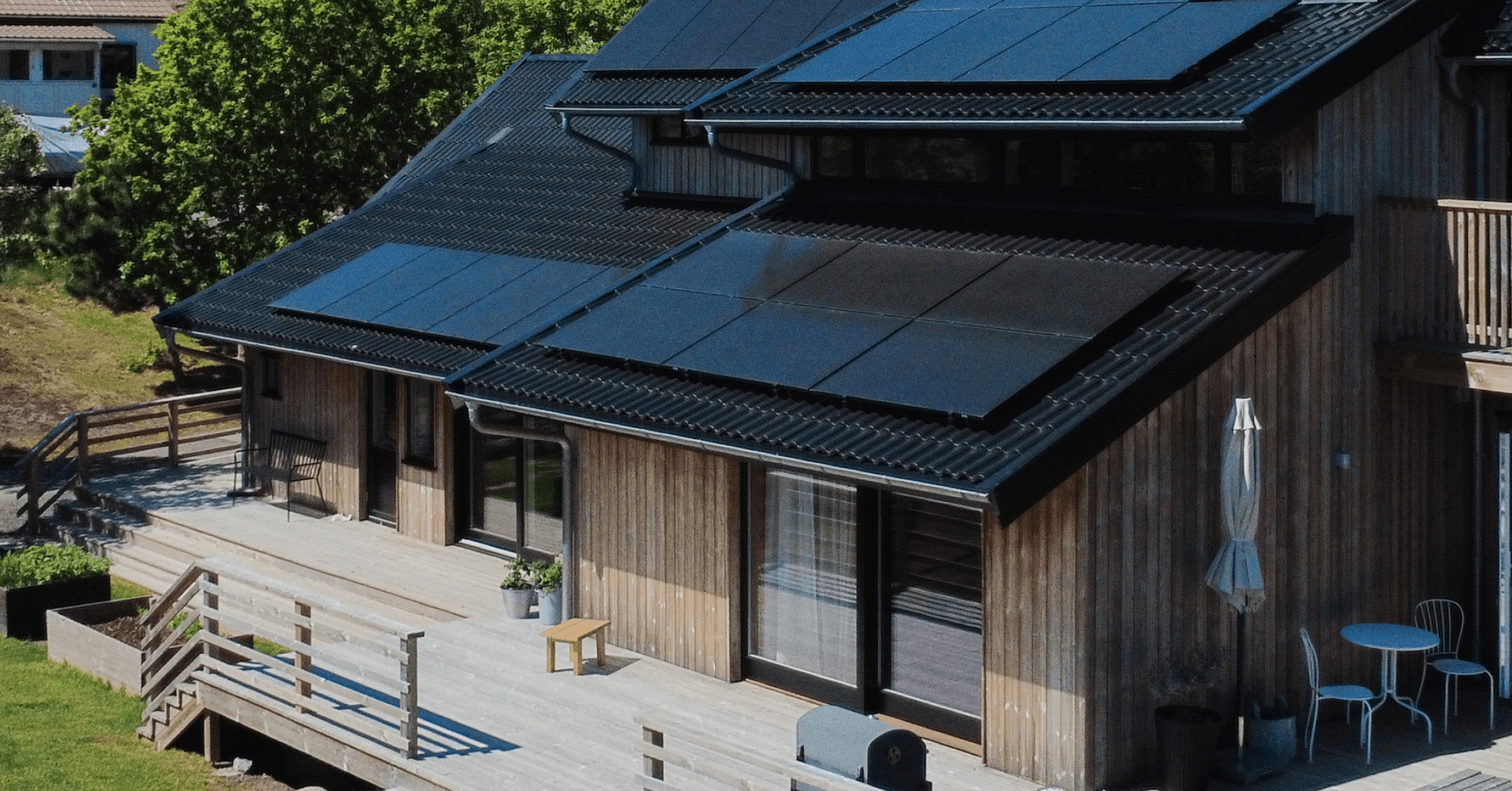 Moderni puutalo, jonka katolla on aurinkopaneelit, tilavalla terassilla ulkokalusteita ja vehreyden ympäröimänä.