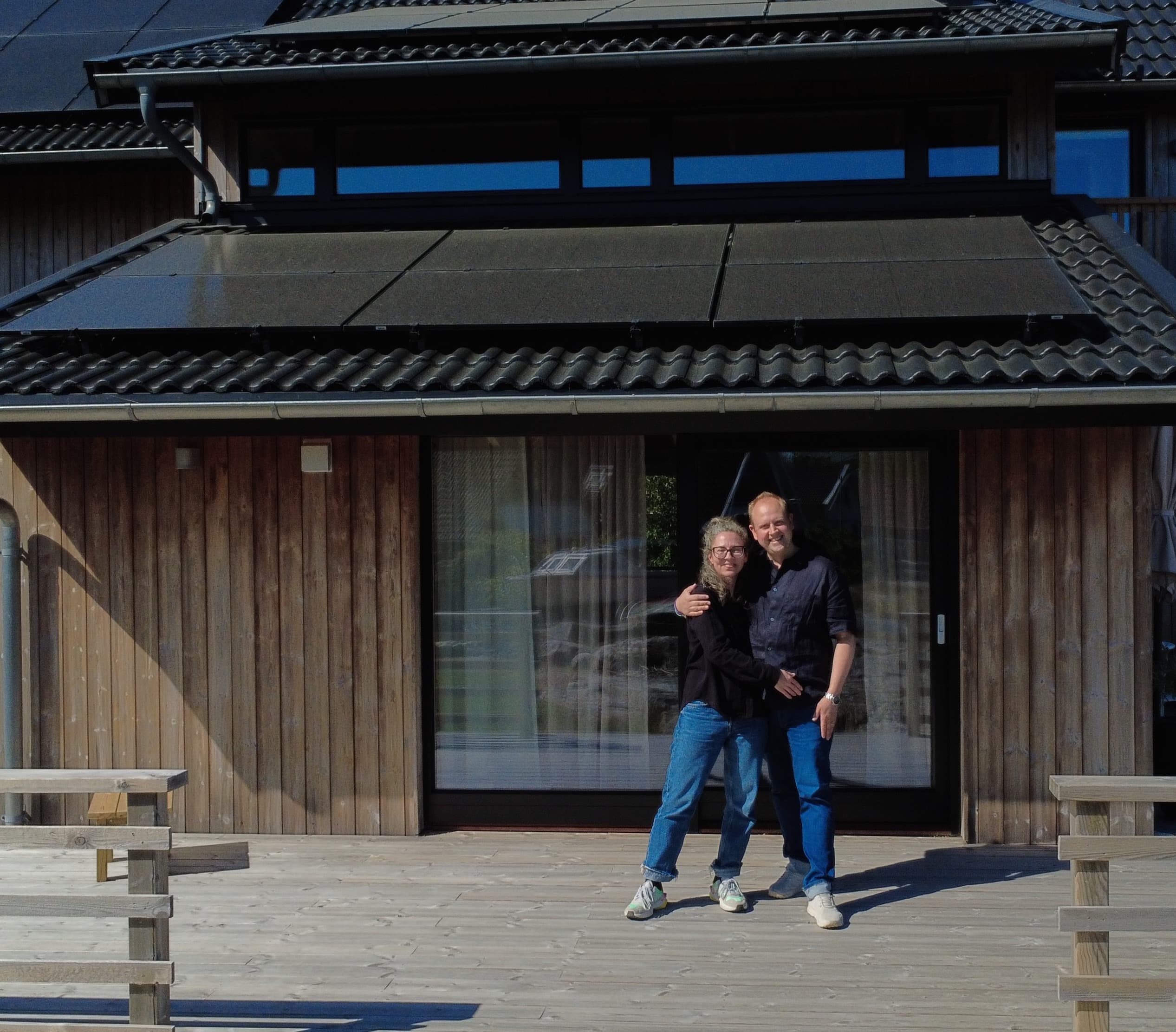 Kaksi ihmistä seisoo hymyillen puisella terassilla modernin puutalon edessä, jossa on suuret ikkunat ja viistokatto.