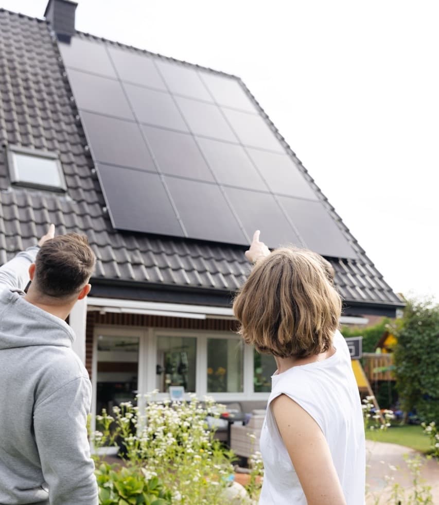 Kaksi ihmistä seisoo talon ulkopuolella, jonka katolla on aurinkopaneelit, ja osoittavat niitä. Talossa on puutarha, jossa kasvaa erilaisia kasveja.