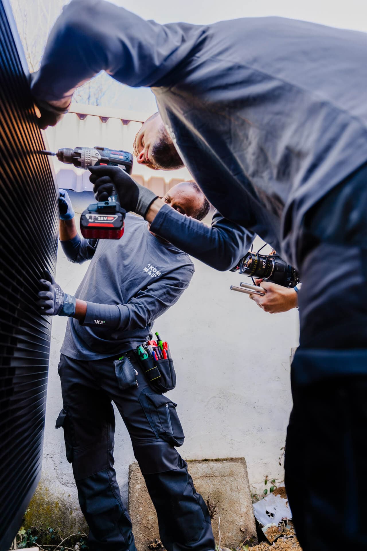 Kaksi työntekijää harmaissa työasuissa asentaa lämpöpumppua käyttäen sähkötyökaluja.