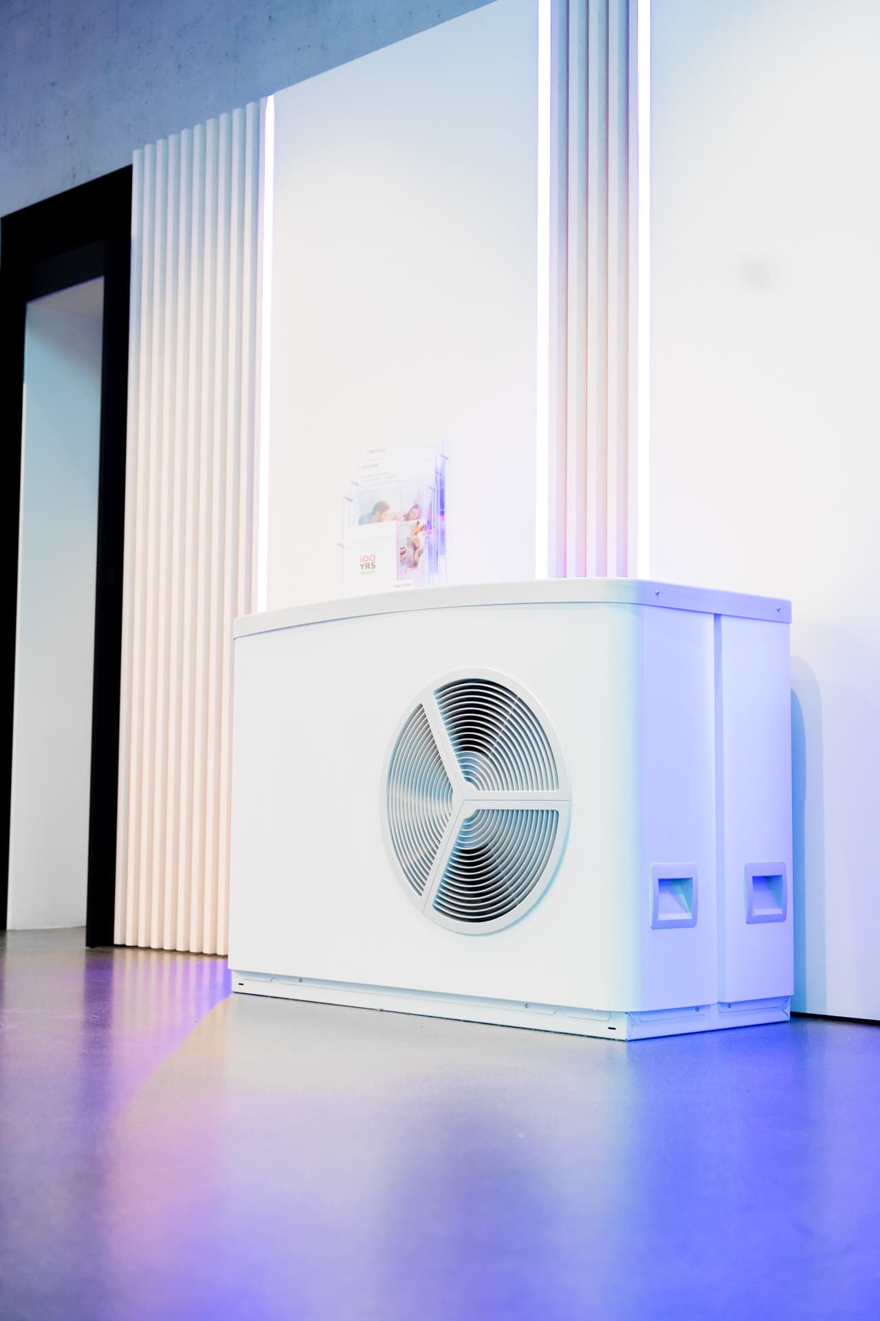 Moderni valkoinen lämpöpumppuyksikkö, jossa on pyöreä ilmanvaihtoaukko, sijoitettuna sisätiloihin seinää vasten, jossa on pystysuuntaisia valonauhoja.