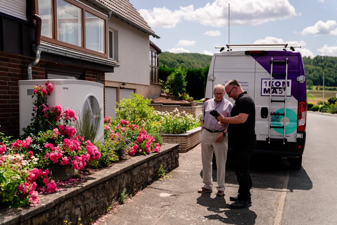 Kaksi miestä seisoo talon vieressä keskustellen valkoisen, violetilla tehosteväreillä varustetun pakettiauton lähellä lämpöpumpusta. Heidän vierellään kukkii kirkkaan pinkkejä kukkia aurinkoiselta taivaalta.