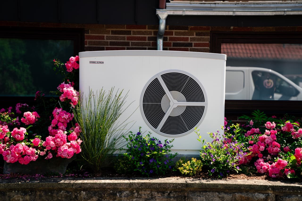 Valkoinen lämpöpumppuyksikkö on värikkäiden pinkkien kukkien ja vehreyden ympäröimä, taustalla tiiliseinä, jossa on ikkuna, ja pakettiauto.