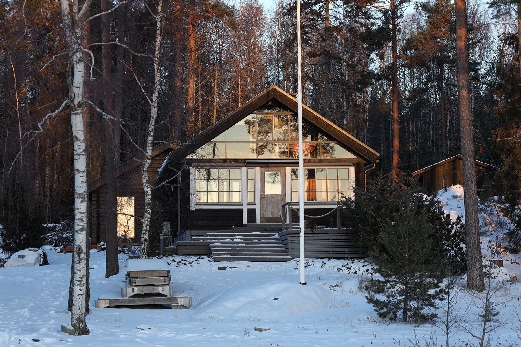 Hirsimökki, jossa on suuret ikkunat, seisoo lumisessa metsässä korkeiden puiden ympäröimänä, edessään lipputanko.