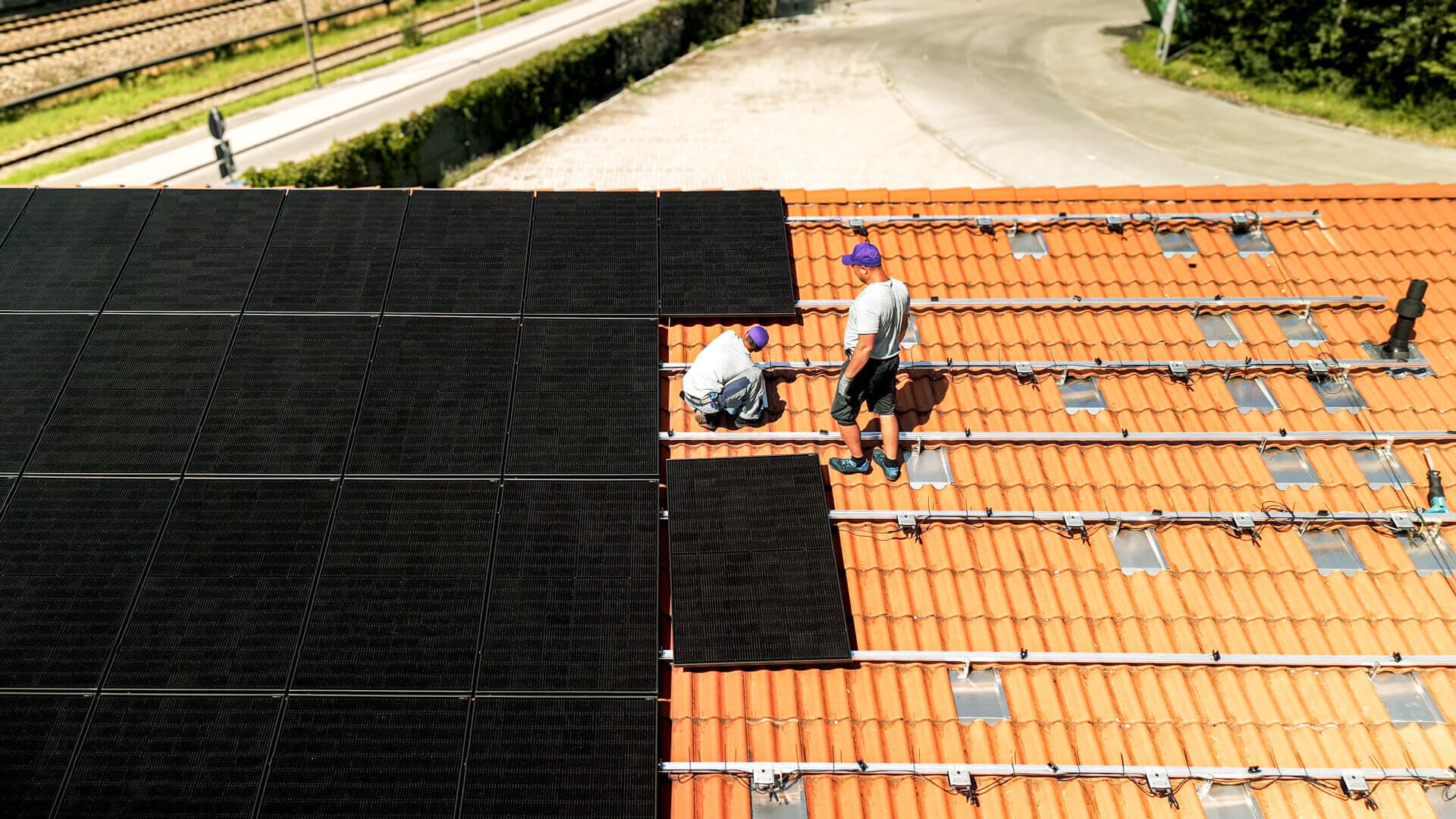 Kaksi asentajaa asentaa aurinkopaneeleita punatiilikatolle tien läheisyydessä, vehreyden ympäröimänä.