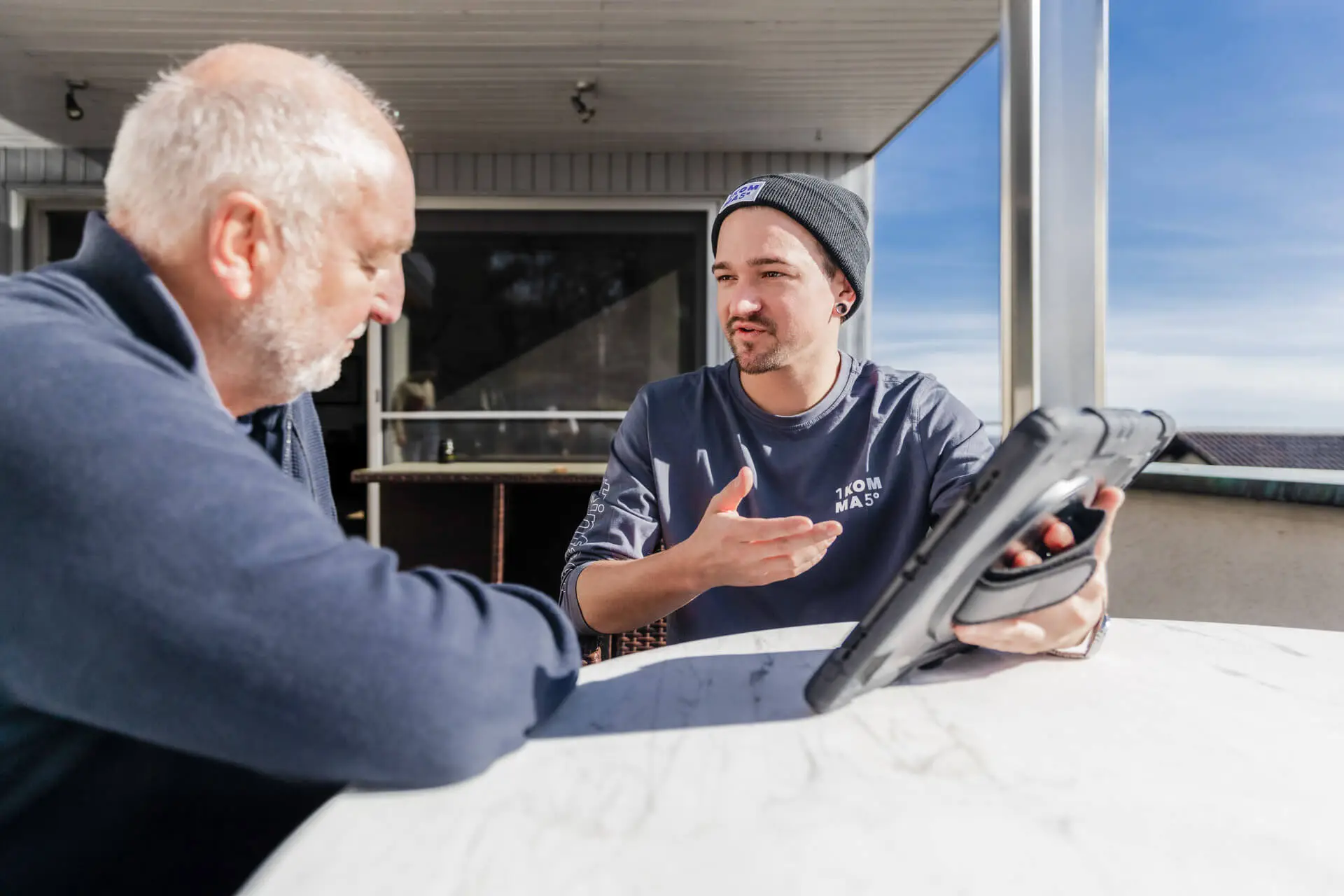 Kaksi miestä istuu ulkona keskustellen. Toinen elehtii kädellään, kun toinen kuuntelee pitäen tablettia kädessään.