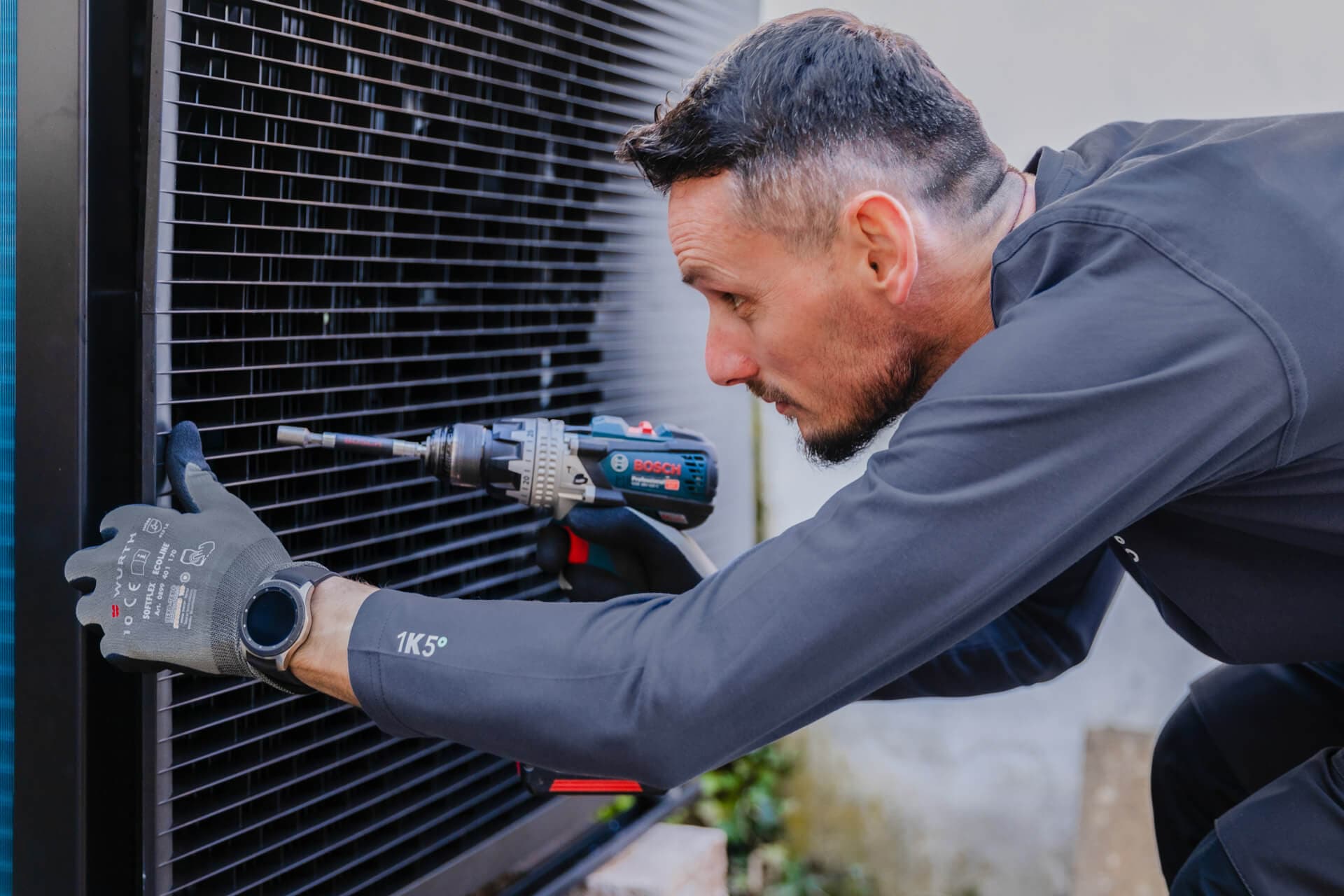 Mies työkkäsineissä käyttää poraa kiinnittäessään tai asentaessaan suurta metalliritilää lämpöpumppuun ulkona.