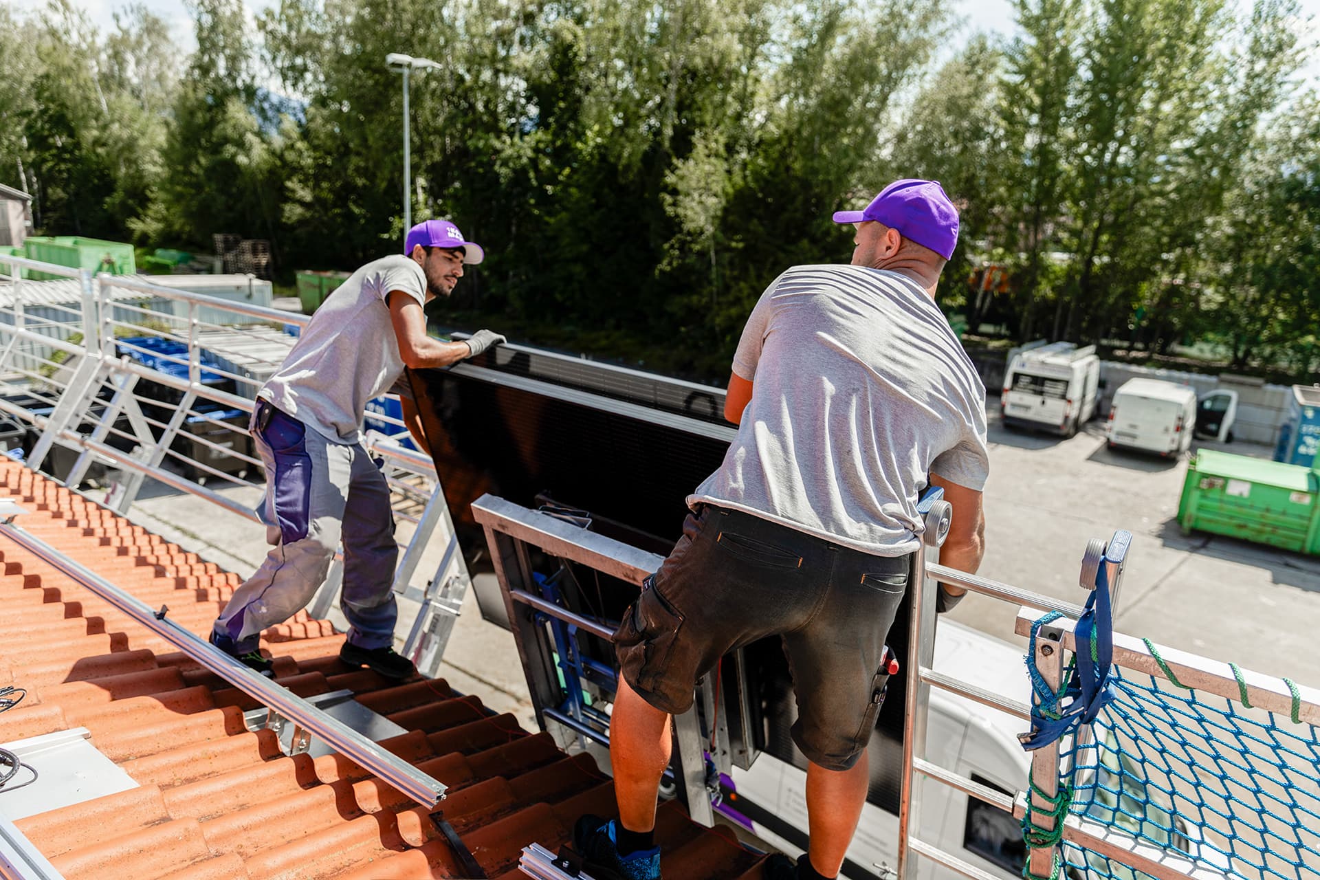 Kaksi työntekijää violetit lippikset päässään asentaa aurinkopaneelia kattolle. Taustalla näkyy puita ja pysäköityjä ajoneuvoja.