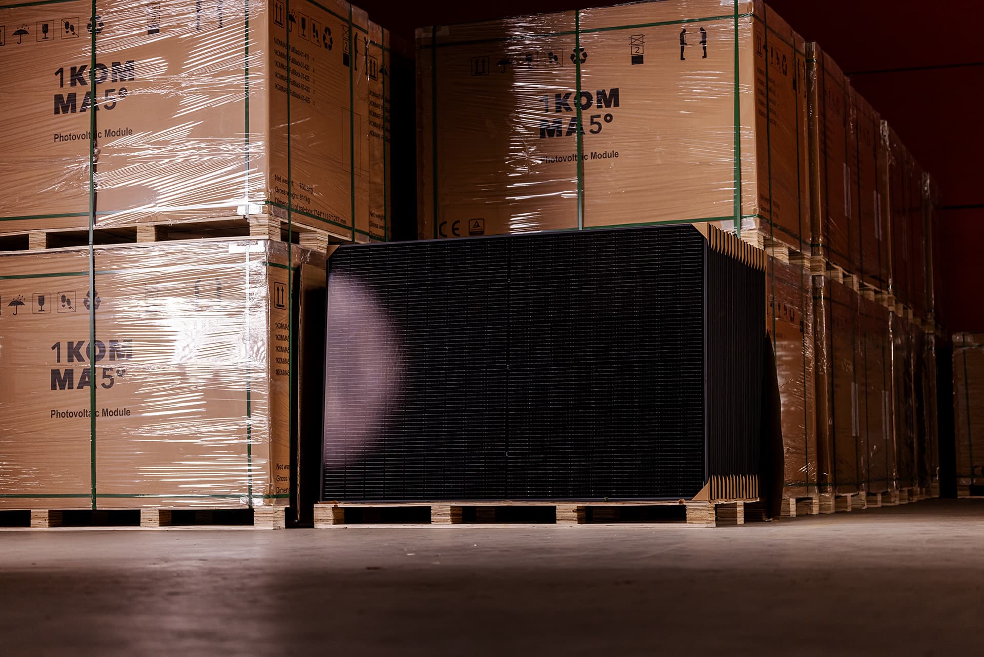 Suuria pahvilaatikkopinoja puisilla lavoilla hämärästi valaistussa varastossa, yhden pinon kylkeen nojaa yksittäinen aurinkopaneeli.