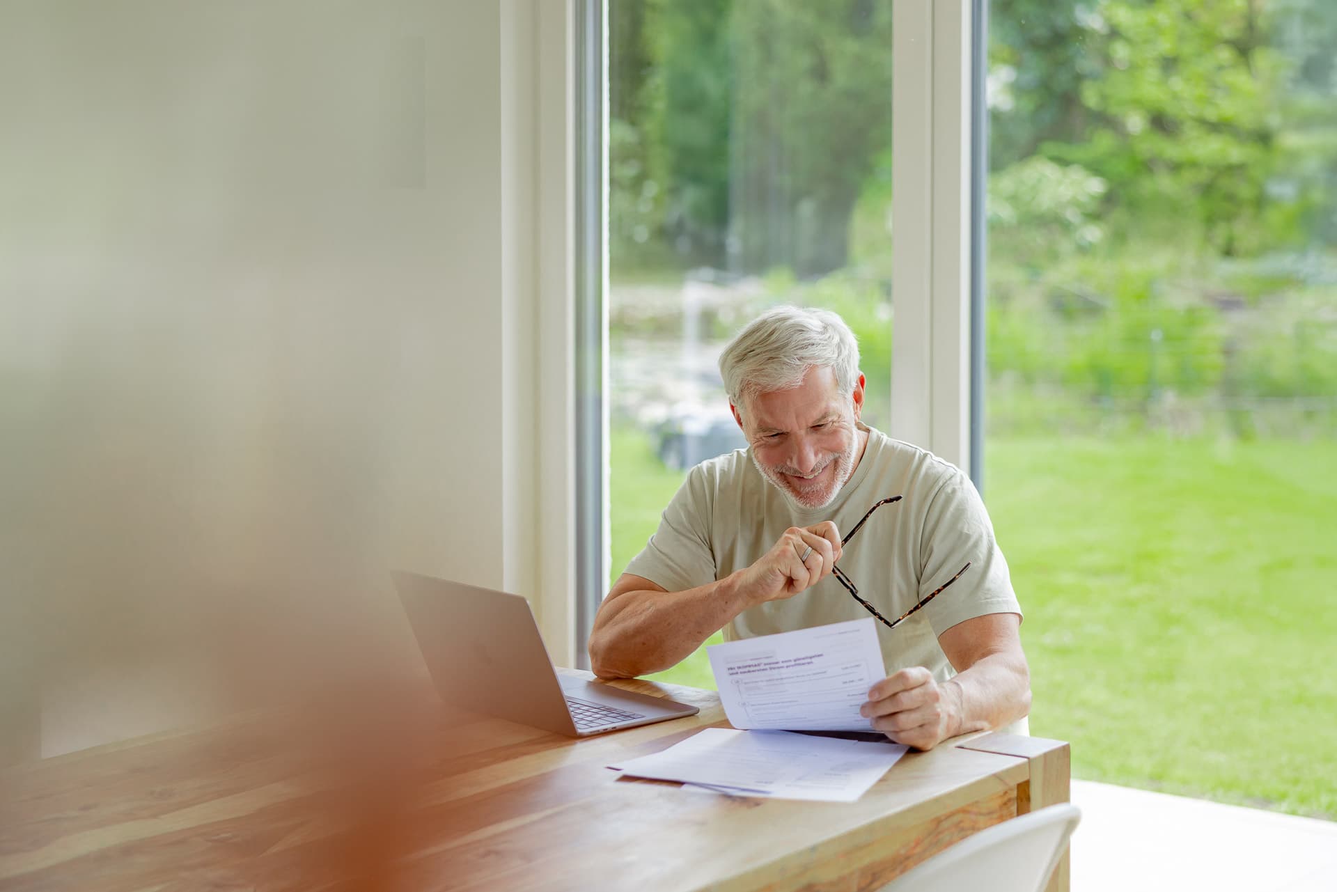 Iäkkäämpi mies hymyilee lukiessaan asiakirjaa puupöydän ääressä, jossa on myös kannettava tietokone, suuren ikkunan vieressä, josta avautuu näkymä puutarhaan.